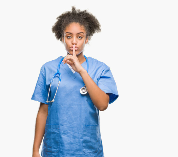 Νέοι afro Αμερικανός γιατρός γυναίκα πέρα από το απομονωμένο υπόβαθρο ζητώντας να είναι ήσυχη με το δάχτυλο στα χείλη. Σιωπή και μυστικό έννοια. - Φωτογραφία, εικόνα