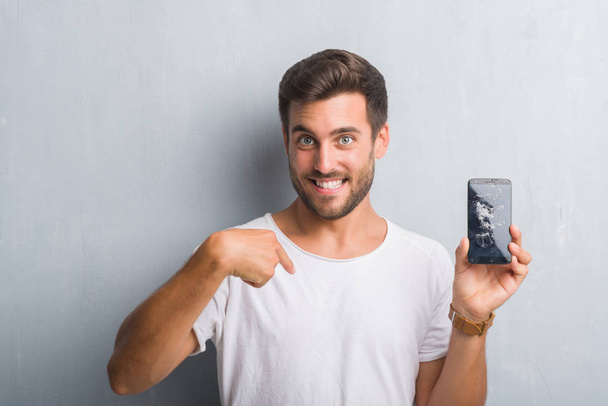 Handsome young man over grey grunge wall showing broken smartphone screen with surprise face pointing finger to himself - Photo, Image