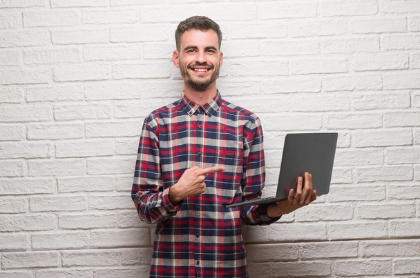 Young adult man over brick wall using laptop very happy pointing with hand and finger - Photo, Image