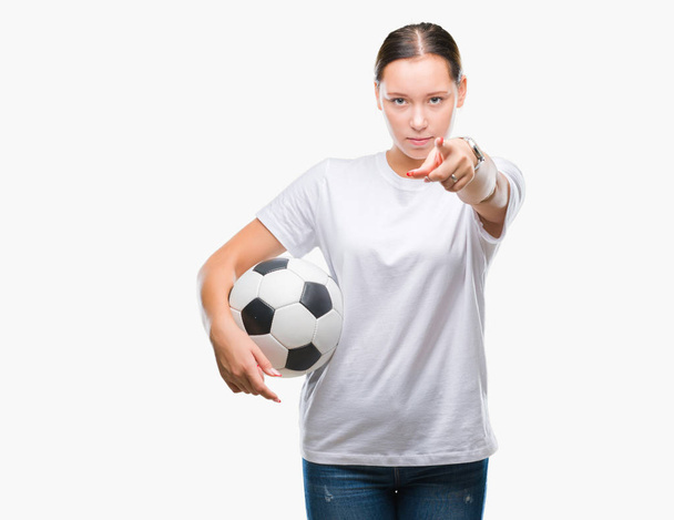 Молодая красивая белая женщина держит футбольный мяч на изолированном фоне, указывая пальцем на камеру и на вас, знак руки, позитивный и уверенный жест спереди
 - Фото, изображение