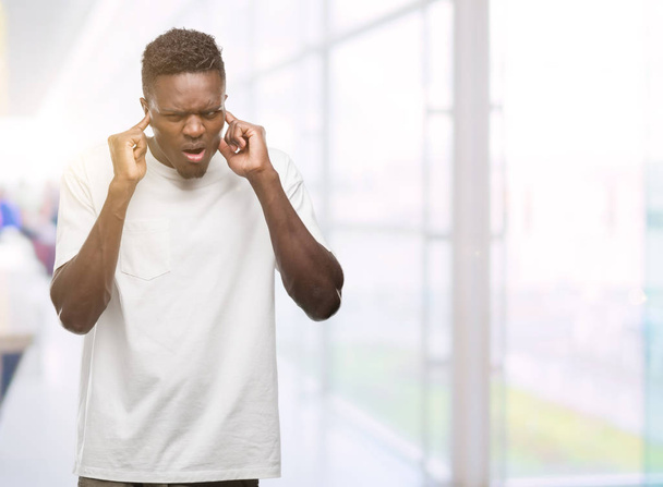 Hombre afroamericano joven con camiseta blanca que cubre las orejas con los dedos con expresión molesta por el ruido de la música alta. Concepto sordo
. - Foto, imagen