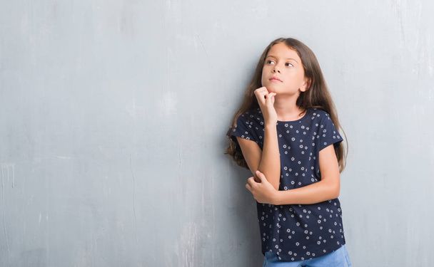 Young hispanic kid over grunge grey wall with hand on chin thinking about question, pensive expression. Smiling with thoughtful face. Doubt concept. - Foto, Imagen