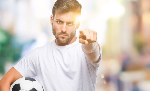 Jovem homem bonito segurando bola de futebol sobre fundo isolado apontando com o dedo para a câmera e para você, sinal de mão, gesto positivo e confiante da frente
 - Foto, Imagem