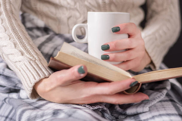 Frau mit Buch und Tasse Kaffee in der Hand trägt Pullover und ist mit einer Retro-Decke bedeckt. Winter- und Kaltwetterkonzept. Nahaufnahme, selektiver Fokus - Foto, Bild