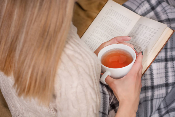 Mujer joven en la cama leyendo un libro y sosteniendo una taza de té. Chica lleva suéter de lana y cubierto con una manta retro. Concepto de clima frío e invierno. Primer plano, enfoque selectivo
 - Foto, Imagen