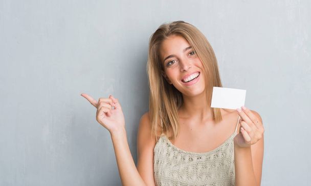 Όμορφη νεαρή γυναίκα πάνω από grunge γκρίζο τοίχο κρατώντας κενό επίσκεψη κάρτα πολύ χαρούμενος, δείχνοντας με το χέρι και δάχτυλο προς την πλευρά - Φωτογραφία, εικόνα