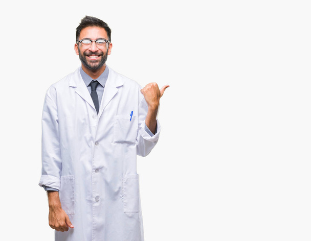 Científico hispano adulto u hombre médico usando una bata blanca sobre un fondo aislado sonriendo con la cara feliz mirando y señalando hacia un lado con el pulgar hacia arriba
. - Foto, imagen