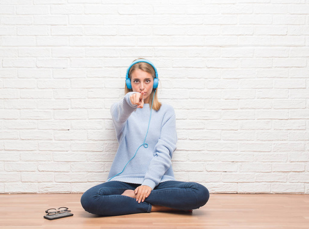 Mulher bonita sentada no chão ouvindo música usando fones de ouvido em casa apontando com o dedo para a câmera e para você, sinal de mão, gesto positivo e confiante da frente
 - Foto, Imagem