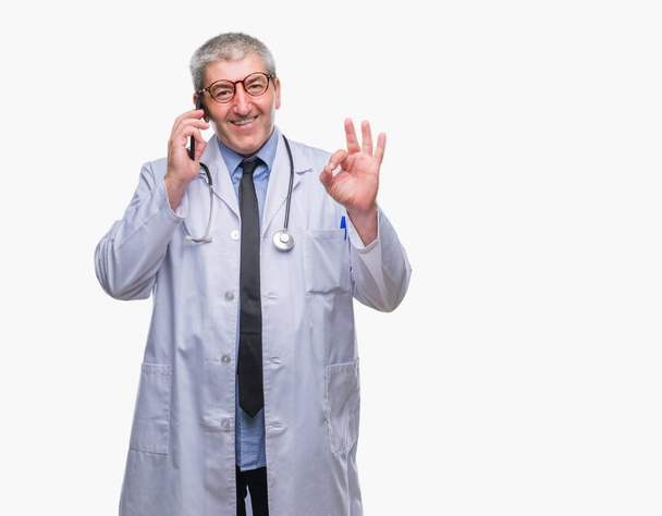 Bonito médico sênior homem falando sobre smarpthone sobre fundo isolado fazendo sinal ok com os dedos, excelente símbolo
 - Foto, Imagem