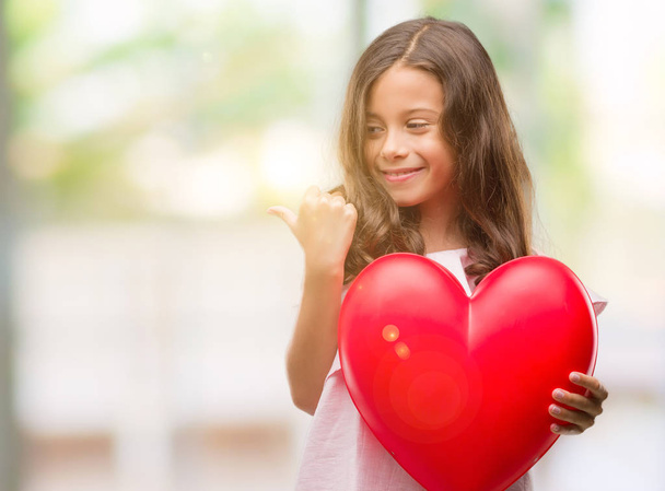 Menina hispânica morena segurando coração vermelho apontando e mostrando com o polegar para o lado com o rosto feliz sorrindo
 - Foto, Imagem