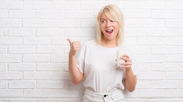 Mujer caucásica adulta sobre pared de ladrillo blanco bebiendo vaso de leche señalando y mostrando con el pulgar hacia el lado con la cara feliz sonriendo
 - Foto, imagen