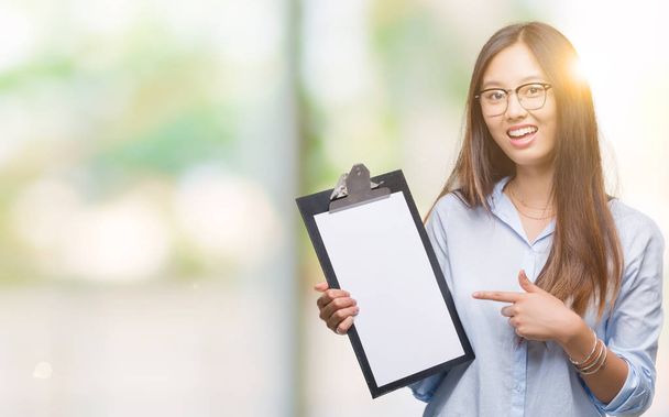 Young asian business woman holding clipboard over isolated background very happy pointing with hand and finger - Фото, изображение