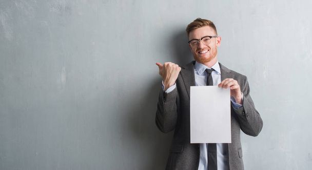 Young redhead  business man over grey grunge wall holding blank paper contract pointing and showing with thumb up to the side with happy face smiling - 写真・画像