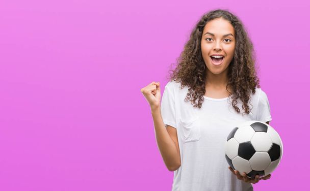 Young hispanic woman holding soccer football ball screaming proud and celebrating victory and success very excited, cheering emotion - Photo, Image