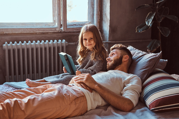 ハンサムな若い父と娘読む童話のベッドの上に横たわっている間一緒に. - 写真・画像