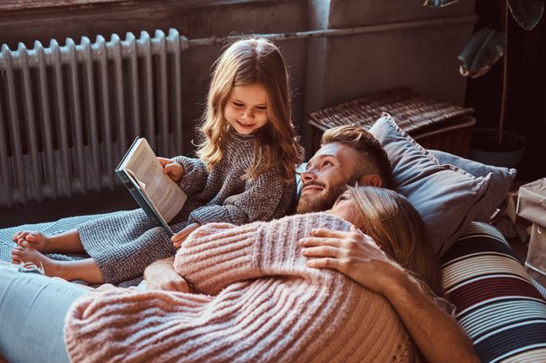 Mom, dad and daughter reading storybook together while lying on bed. - Photo, Image