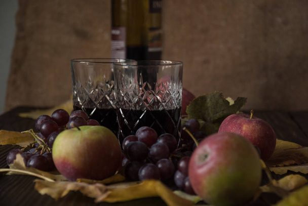 Two crystal glasses with red wine, apples, grapes and dry leaves on a wooden surface - Fotó, kép