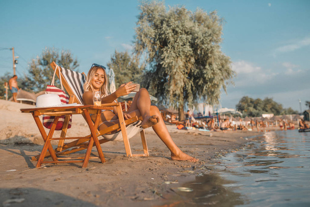 attraktive junge Frau im Badeanzug mit Handtuch auf dem Kopf, die auf einem Liegestuhl am Strand sitzt und ein Selfie mit dem Handy macht  - Foto, Bild