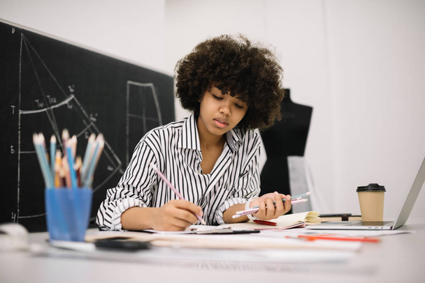 Beautiful African American woman freelancer sketching or drawing at workplace. Portrait of cute stylish curly hair fashion illustrator in modern office or atelier. Successful business concept.  - Fotó, kép