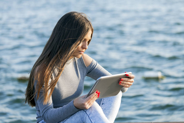 Mujer joven adolescente sentado junto al océano / río y el uso de la tableta. Mujer utilizar tableta táctil PC tecnología de Internet. Copiar espacio
 - Foto, Imagen