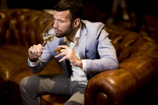 Rich young businessman tasting white wine and smoking cigar - Foto, Imagen