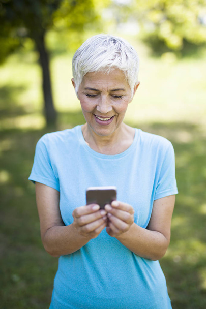 Portret sportowy senior kobiety przy użyciu telefonu komórkowego w parku - Zdjęcie, obraz