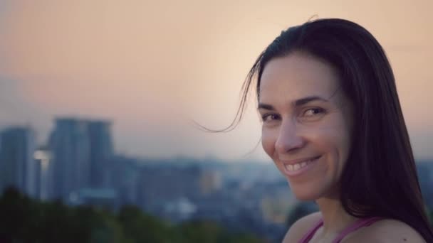 Şehrin arka plan üzerinde çekici bir sporcumuz portresi. Sportif bir yapı olan sevimli kadın kameraya bakıyor. Uzun saçlı esmer günbatımı uzakta fonunda poz - Video, Çekim