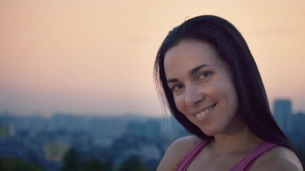 Portrait of an attractive sportswoman on the background of the city. Cute young woman with a sporty build is looking at the camera. Long-haired brunette posing against the backdrop of the sunset away - Footage, Video