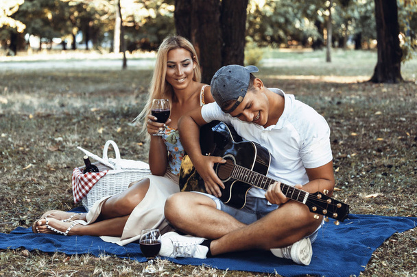 Young man playing guitar to his girl summer day couple love sitting green grass outdoor picnic nature guy singing songl. They enjoy nature - Fotografie, Obrázek