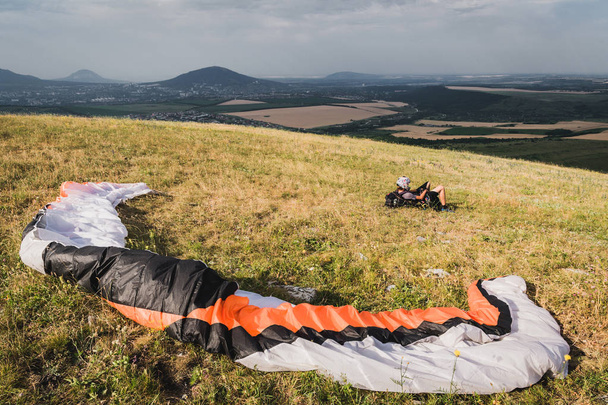 A professional paraglider in full gear and a helmet lies and rests on the grass high in the mountains looking at the clouds - Fotó, kép