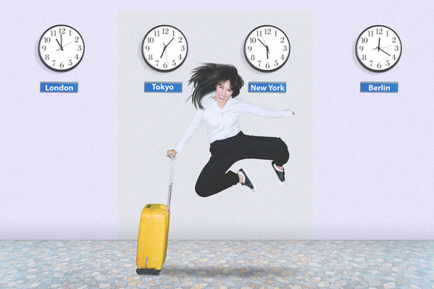 Happy female entrepreneur jumping in the airport while holding a luggage. Shot with different country clock's background - Fotografie, Obrázek