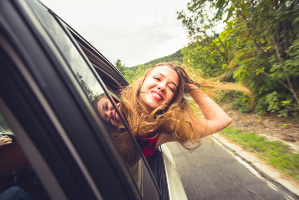 Uma menina está com a cabeça para fora de uma janela do carro enquanto está dirigindo na natureza, sorrindo e se divertindo
 - Foto, Imagem