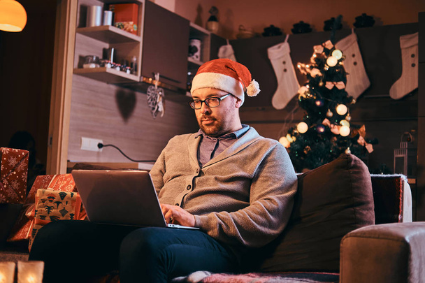 Man in Santa hat and glasses dressed in warm sweater working on laptop at Christmas. - Photo, Image