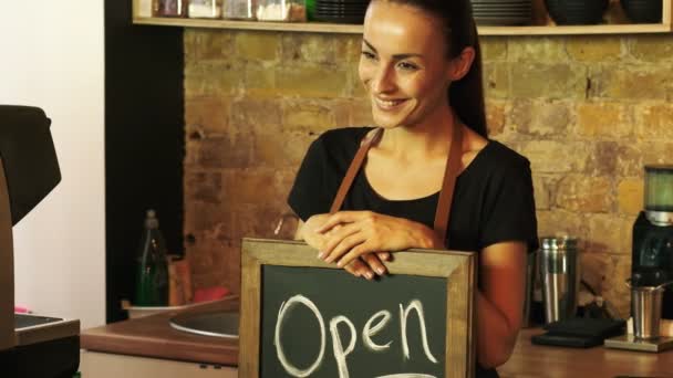 Un trabajador de una cafetería sostiene un letrero abierto y mira al cliente
. - Imágenes, Vídeo