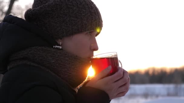mooi meisje drinkt hete thee uit haar beker glas bij zonsondergang, in het park in de winter. meisje verwarmt haar handen op glas koffie in het winter forest - Video