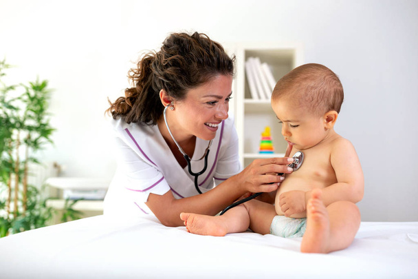 Physician examining her baby patient with stethoscope as the baby sits upright during medical examination - Zdjęcie, obraz