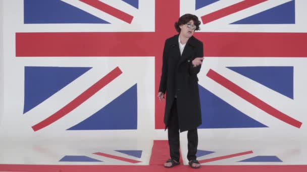 Смешной парень в парике, солнечных очках и пальто говорит на камеру в студии с флагом Великобритании
 - Кадры, видео