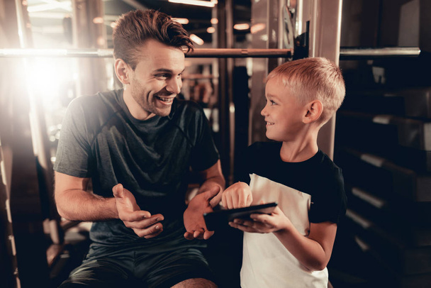 Father And Son In Gym. Information On Tablet. Parenthood Relationship. Sporty Family Concept. Active Lifestyle. Happy Childhood. Holiday Leisure. Working Out Together. Fitness Day. - Zdjęcie, obraz