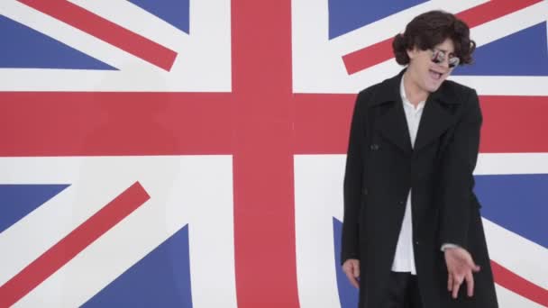 Молодой человек в парике, солнцезащитных очках и пальто поет на камеру в студии с флагом Великобритании
 - Кадры, видео