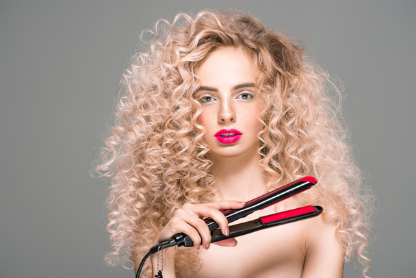 beautiful naked girl with long curly hair holding hair straightener and looking at camera isolated on grey - 写真・画像