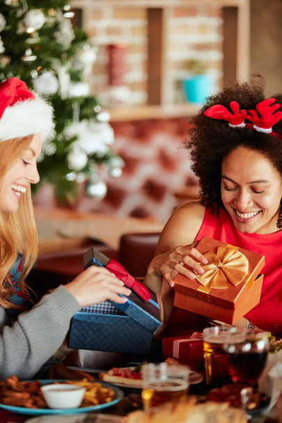 Vrienden geven van geschenken aan elkaar zittend aan tafel. In achtergrond kerstboom. Kerst vakantie concept. - Foto, afbeelding