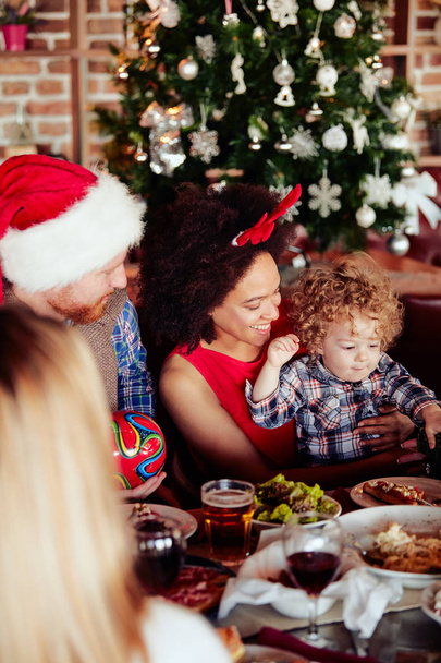 お友達とテーブルに座って幼児にギフトを与えます。背景のクリスマス ツリー。クリスマス休暇の概念. - 写真・画像