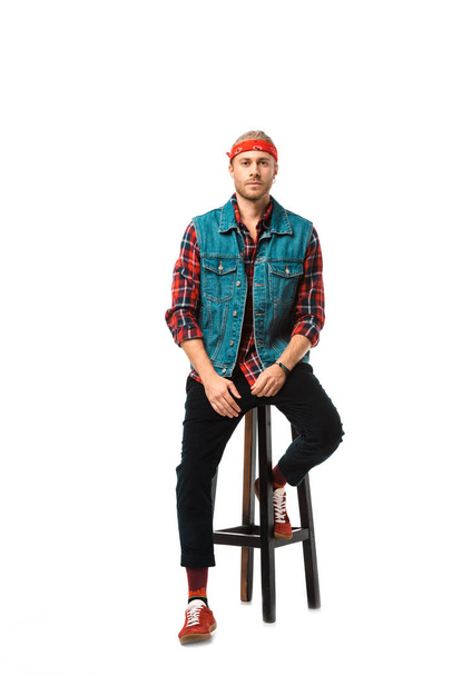modische Hipster-Mann in Jeansweste und rot kariertem Hemd sitzt auf Stuhl isoliert auf weiß  - Foto, Bild