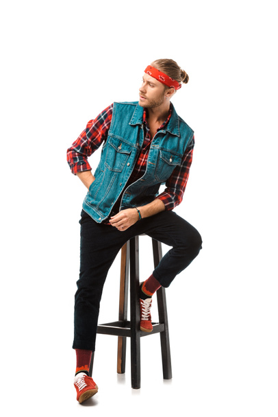 young hipster man in denim vest and red checkered shirt posing on chair isolated on white  - Foto, afbeelding