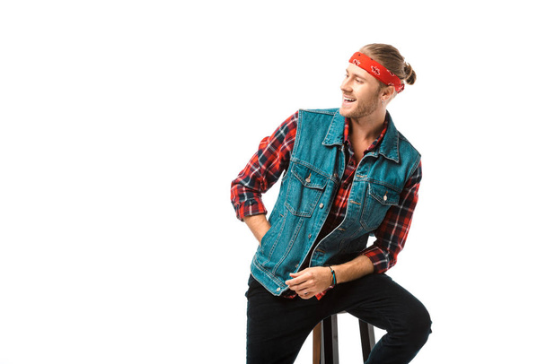 happy hipster man in denim vest and red checkered shirt looking away on chair isolated on white  - Fotoğraf, Görsel