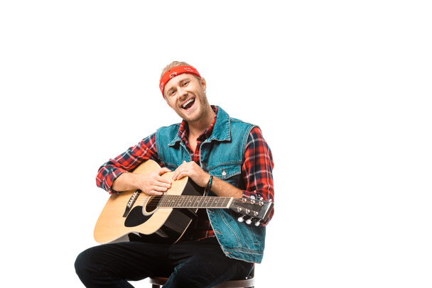 smiling hipster man in denim vest sitting with acoustic guitar isolated on white - Fotó, kép