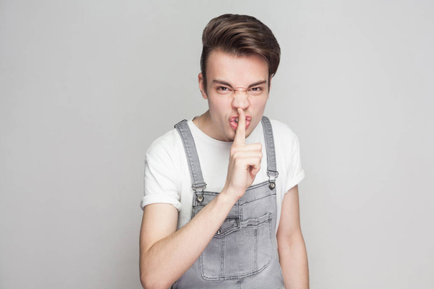 Portrait of angry young brunette man in casual style with white t-shirt and denim overalls standing and looking at camera while showing quiet sign on gray background. - Фото, изображение