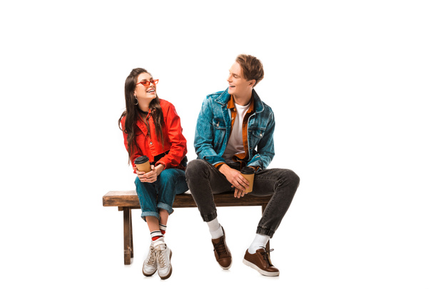 laughing hipster couple with disposable cups of coffee sitting on bench isolated on white - Foto, afbeelding