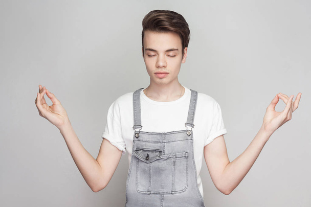Portrait of calm relax young brunette man in casual style with white t-shirt and denim overalls standing with closed eyes and doing yoga training on gray background. - Photo, image