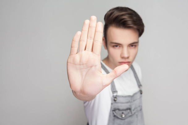 Serious young brunette man in casual style with white t-shirt and denim overalls looking at camera and showing stop gesture on gray background. - Fotografie, Obrázek